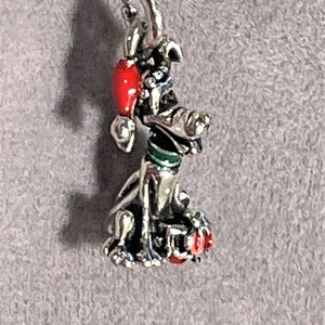 May include: Silver charm featuring a cartoon dog wearing a red Santa hat with green trim. The dog is sitting with its head tilted to the side and its tongue sticking out.