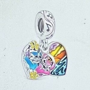 May include: A silver charm with a heart-shaped pendant featuring a colorful design. The pendant showcases a blue and pink cartoon character embracing, with a rainbow and musical notes. Yellow flower accents add detail.