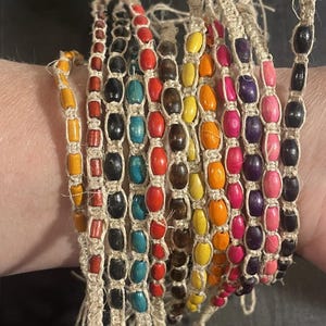 May include: A collection of colorful beaded bracelets. Each bracelet features wooden beads in various colors, including red, yellow, blue, and black, strung on natural twine. The bracelets are stacked together, showcasing a vibrant and bohemian style.