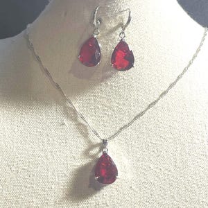 May include: A jewelry set consisting of a silver-toned chain necklace and a pair of matching drop earrings. Each piece features a faceted, teardrop-shaped red gemstone set in a silver-toned metal frame. The necklace has a delicate twisted chain.