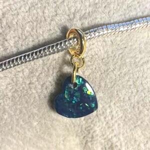 May include: A gold-colored charm with a dark blue heart-shaped pendant. The pendant has a shimmering, iridescent effect with green and yellow accents. The charm is attached to a silver chain.