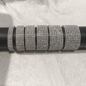 May include: A collection of silver rhinestone bracelets displayed on a black cylindrical stand. The bracelets vary in width, each covered in sparkling, clear rhinestones. The background is a soft, white fabric.
