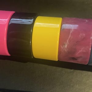 May include: A collection of five colorful, wide, glossy bangles. The colors include hot pink, black, yellow, marbled pink, and navy blue. These bold, chunky bracelets are a statement accessory.