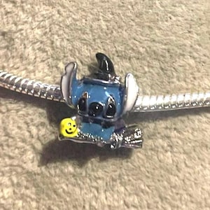 K&ouml;nnte beinhalten: Ein blauer und schwarzer Stitch-Charm mit wei&szlig;en Ohren und einem Hexenhut. Der Charm h&auml;lt ein gelbes Smiley-Gesicht und ist an einem silbernen Armband befestigt. Der Charm ist ein Accessoire im Disney-Stil.