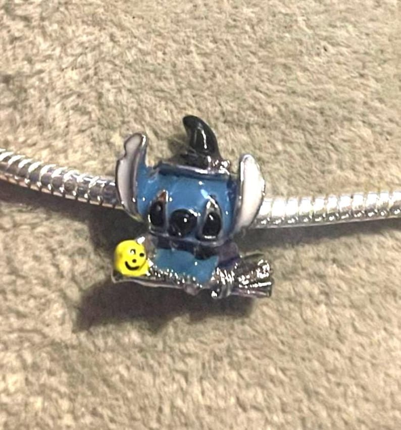 K&ouml;nnte beinhalten: Ein blauer und schwarzer Stitch-Charm mit wei&szlig;en Ohren und einem Hexenhut. Der Charm h&auml;lt ein gelbes Smiley-Gesicht und ist an einem silbernen Armband befestigt. Der Charm ist ein Accessoire im Disney-Stil.