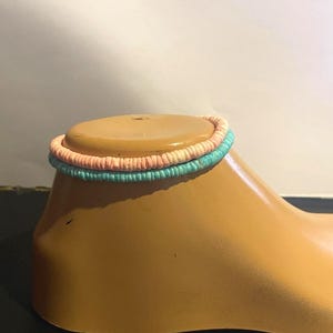 May include: A stack of two colorful bracelets. The top bracelet is a light peach color, and the bottom bracelet is a light turquoise color. The bracelets are made of a woven material.