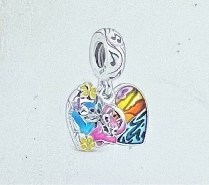 May include: A silver charm with a heart-shaped pendant featuring a colorful design. The pendant showcases a blue and pink cartoon character embracing, with a rainbow and musical notes. Yellow flower accents add detail.