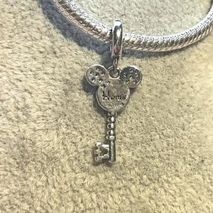 May include: A silver charm in the shape of a key, with a Mickey Mouse head design at the top. The word "Home" is inscribed on the charm. The charm is attached to a silver bracelet.