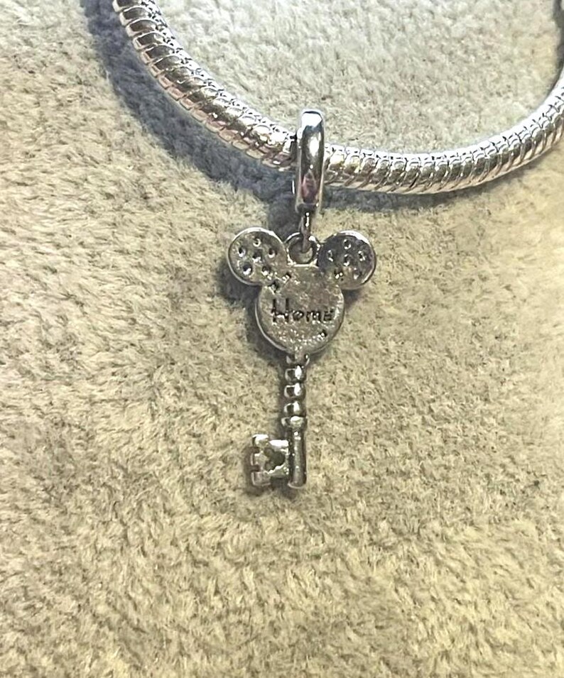 May include: A silver charm in the shape of a key, with a Mickey Mouse head design at the top. The word "Home" is inscribed on the charm. The charm is attached to a silver bracelet.