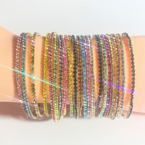 May include: A collection of colorful, thin bangles with iridescent rhinestones. The bangles are stacked on a light-toned arm, showcasing a variety of colors including pink, green, yellow, and blue. The jewelry sparkles in the light.