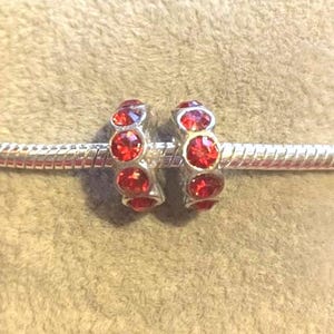 May include: Two silver-toned charm beads with a row of three red gemstones. The beads are on a silver bracelet. The gemstones are round and faceted, reflecting light. The background is a soft, neutral color.