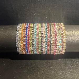 May include: A multi-colored rhinestone bracelet with vertical stripes of various colors, including orange, blue, green, and pink. The bracelet is set on a black surface, showcasing its sparkling details.