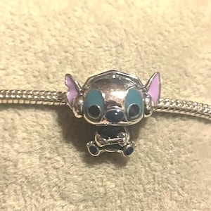 May include: A silver charm featuring the character Stitch from the Disney movie Lilo & Stitch. The charm has blue eyes, purple ears, and a dark blue nose. The charm is attached to a silver bracelet.