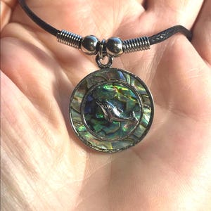 May include: A round pendant necklace featuring a silver dolphin charm set against an iridescent abalone shell background. The pendant is suspended from a black cord necklace with silver-toned metal accents.