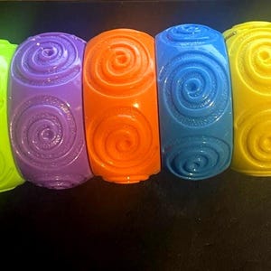 May include: A collection of six colorful bangles in neon green, purple, orange, blue, yellow, and pink. Each bangle features a raised spiral design. These bold, chunky bracelets are a vibrant accessory.