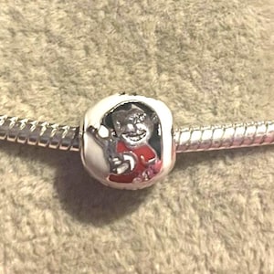 Pode incluir: Um berloque de prata com um personagem de desenho animado com uma camisa vermelha e uma borda branca. O berloque est&aacute; em uma corrente de prata, em um fundo de tecido marrom claro texturizado.