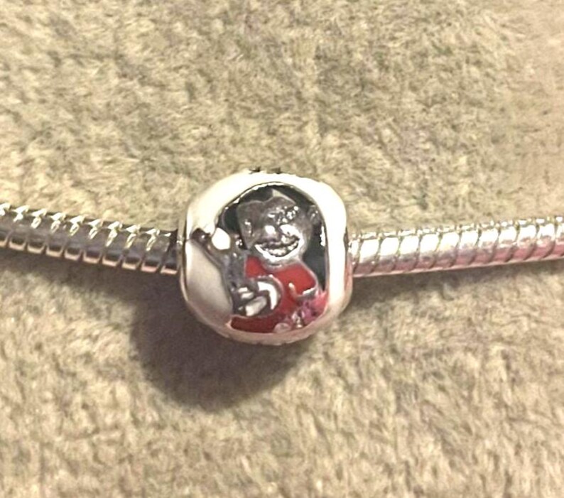 Pode incluir: Um berloque de prata com um personagem de desenho animado com uma camisa vermelha e uma borda branca. O berloque est&aacute; em uma corrente de prata, em um fundo de tecido marrom claro texturizado.