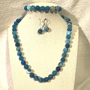 May include: A three-piece jewelry set featuring a necklace, bracelet, and earrings. The set is made of blue and white striped beads. The necklace and bracelet are strung with round beads, while the earrings have a single bead each.