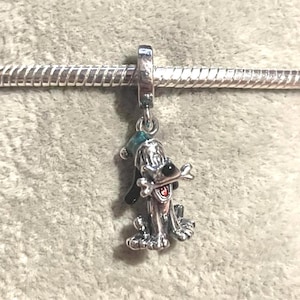 May include: A silver-colored charm featuring a cartoon dog holding a bone in its mouth. The dog has black ears and a green hat. The charm is attached to a silver chain.