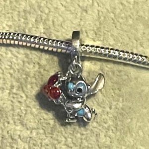 Pode incluir: Um berloque de prata com o personagem Stitch de Lilo & Stitch. Stitch &eacute; retratado com pele azul, olhos grandes e pretos e um objeto vermelho em forma de cora&ccedil;&atilde;o. O berloque est&aacute; preso a uma corrente de prata.