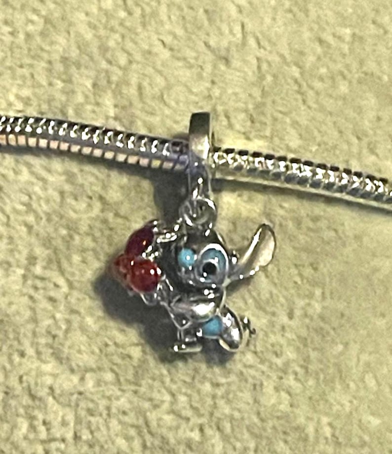 Pode incluir: Um berloque de prata com o personagem Stitch de Lilo & Stitch. Stitch &eacute; retratado com pele azul, olhos grandes e pretos e um objeto vermelho em forma de cora&ccedil;&atilde;o. O berloque est&aacute; preso a uma corrente de prata.