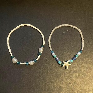 May include: Two beaded anklets. One features white beads with pearl and teal accents. The other has white beads, teal and blue accents, and a white starfish charm. Both are on a dark surface.