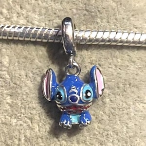Puede incluir: Un charm azul y plateado con un personaje de dibujos animados con orejas grandes y una amplia sonrisa. El charm cuelga de una cadena plateada, mostrando las caracter&iacute;sticas distintivas y los colores vibrantes del personaje.