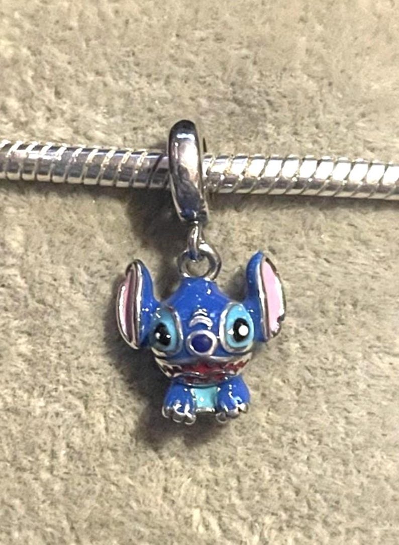 Puede incluir: Un charm azul y plateado con un personaje de dibujos animados con orejas grandes y una amplia sonrisa. El charm cuelga de una cadena plateada, mostrando las caracter&iacute;sticas distintivas y los colores vibrantes del personaje.