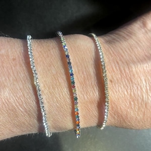 May include: Three delicate bracelets are displayed on a wrist. One is silver with clear stones, another is silver with a rainbow of colored stones, and the third is gold with clear stones. The bracelets are thin and elegant.
