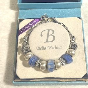 May include: A silver charm bracelet with blue and white beads, white faux pearls, and decorative silver accents. The bracelet is displayed in a blue box with the text "Bella Perlina" and "One size fits all".