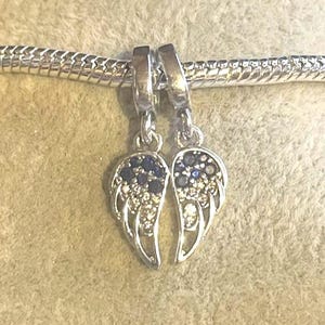 May include: Silver-toned charm featuring two wing pendants. Each wing is adorned with small, sparkling clear and dark blue gemstones. The charms are suspended from a silver chain, creating a delicate and elegant accessory.