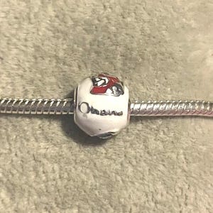 Pode incluir: Uma conta de berloque branca com um design vermelho e preto e a palavra "Ohio" em letras prateadas. O berloque est&aacute; em uma pulseira prateada. A conta &eacute; um elemento decorativo para pulseiras.