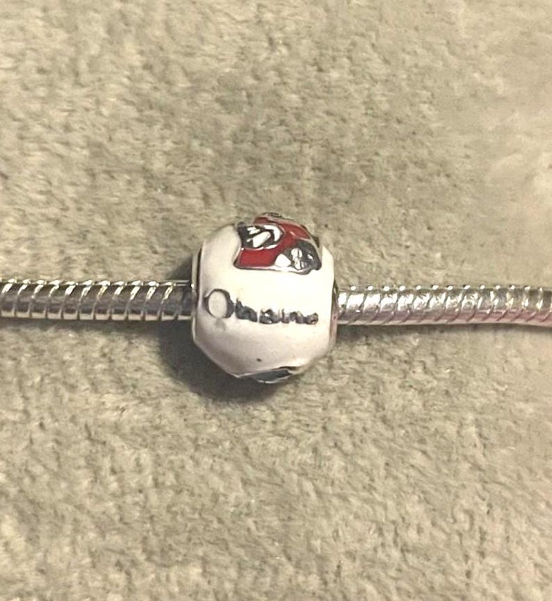 Pode incluir: Uma conta de berloque branca com um design vermelho e preto e a palavra "Ohio" em letras prateadas. O berloque est&aacute; em uma pulseira prateada. A conta &eacute; um elemento decorativo para pulseiras.