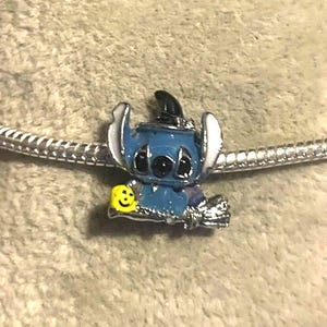 K&ouml;nnte beinhalten: Ein blau-silberner Charm-Anh&auml;nger mit einer Cartoon-Figur, die einen Hexenhut tr&auml;gt und ein gelbes Smiley-Gesicht h&auml;lt. Der Anh&auml;nger befindet sich an einer silbernen Kette, ideal f&uuml;r ein Armband oder eine Halskette.