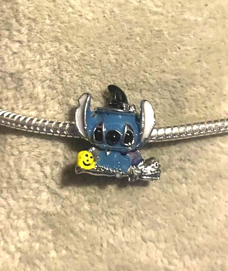 K&ouml;nnte beinhalten: Ein blau-silberner Charm-Anh&auml;nger mit einer Cartoon-Figur, die einen Hexenhut tr&auml;gt und ein gelbes Smiley-Gesicht h&auml;lt. Der Anh&auml;nger befindet sich an einer silbernen Kette, ideal f&uuml;r ein Armband oder eine Halskette.