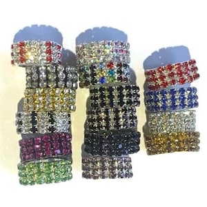 May include: A collection of rhinestone rings in various colors, including red, silver, gold, and green. Each ring is adorned with multiple rows of sparkling rhinestones, creating a dazzling effect. The rings are stacked, showcasing their different designs.