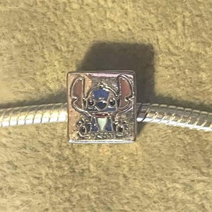 May include: A silver charm featuring the character Stitch from Lilo & Stitch. The charm is square-shaped with a detailed, colorful image of Stitch in blue, pink, and white. The charm is on a silver chain, set against a textured, light brown background.