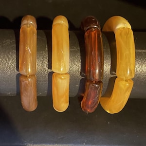 May include: Four vintage bracelets in shades of amber, brown, and yellow. The bracelets are made of cylindrical beads strung together. The colors vary from light to dark, creating a gradient effect.