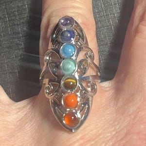 May include: Silver-toned ring featuring seven colorful gemstones in a vertical arrangement. The gemstones include purple, blue, turquoise, yellow-brown, orange, and red. The ring has an ornate, openwork design.