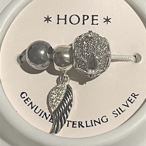 May include: A silver charm bracelet with three charms: a round bead, a charm with a flower design, and a charm with a wing design. The charms are on a white cord. The bracelet is on a white card with the words "HOPE" and "GENUINE STERLING SILVER".