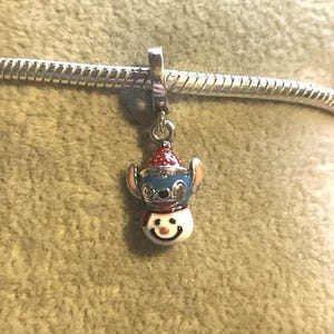 Puede incluir: Un charm con un dise&ntilde;o de mu&ntilde;eco de nieve con cabeza azul, gorro rojo y cara sonriente. El charm est&aacute; sujeto a una pulsera plateada. El charm de mu&ntilde;eco de nieve es un accesorio festivo.