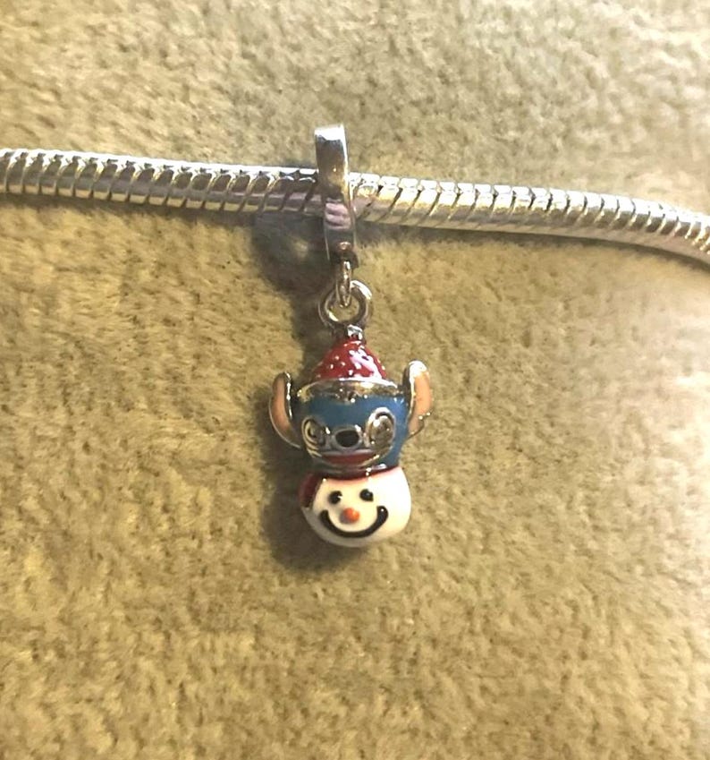 Puede incluir: Un charm con un dise&ntilde;o de mu&ntilde;eco de nieve con cabeza azul, gorro rojo y cara sonriente. El charm est&aacute; sujeto a una pulsera plateada. El charm de mu&ntilde;eco de nieve es un accesorio festivo.