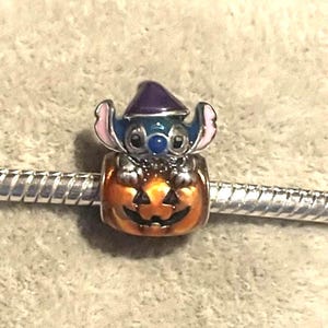 Peut inclure: Un charm repr&eacute;sentant un personnage bleu et rose coiff&eacute; d'un chapeau de sorci&egrave;re violet, assis dans une citrouille orange. Le charm est fix&eacute; &agrave; un bracelet argent&eacute;. Ce charm fait probablement partie d'une collection de bijoux sur le th&egrave;me d'Halloween ou de Disney.