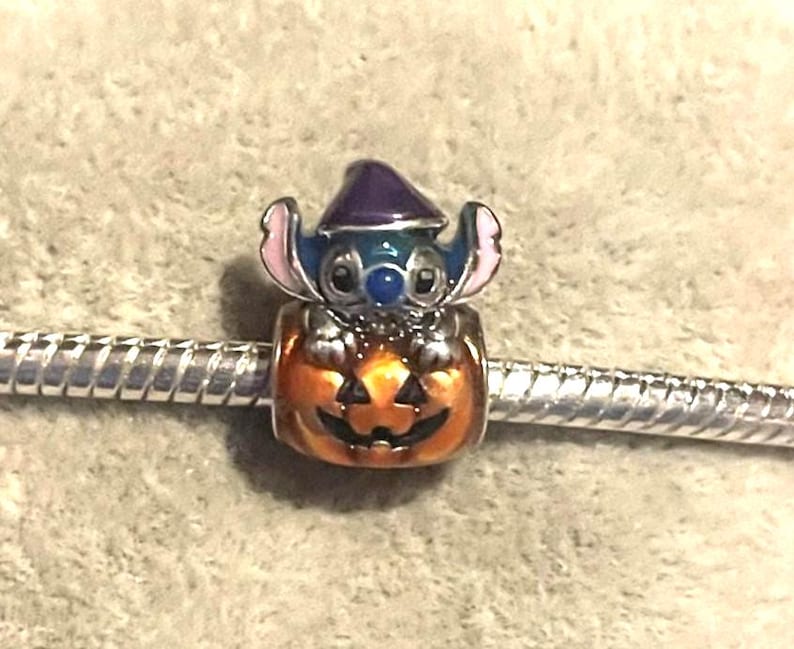 Peut inclure: Un charm repr&eacute;sentant un personnage bleu et rose coiff&eacute; d'un chapeau de sorci&egrave;re violet, assis dans une citrouille orange. Le charm est fix&eacute; &agrave; un bracelet argent&eacute;. Ce charm fait probablement partie d'une collection de bijoux sur le th&egrave;me d'Halloween ou de Disney.