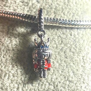 May include: A silver charm depicting a nutcracker soldier with a crown. The charm has red, blue, and green accents. The charm is hanging from a silver chain.