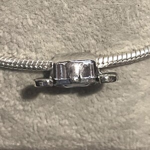 Może przedstawiać: Srebrny charms z prostokątnym środkiem i dwoma małymi, zaokrąglonymi przedłużeniami po bokach. Charms jest przymocowany do srebrnej bransoletki z łańcuszkiem wężowym. Charms ma błyszczącą, odblaskową powierzchnię.