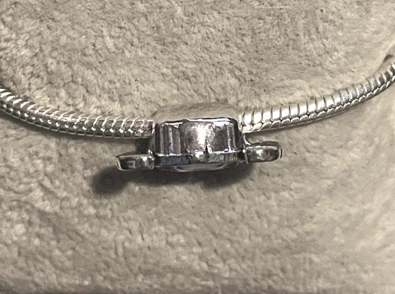 Może przedstawiać: Srebrny charms z prostokątnym środkiem i dwoma małymi, zaokrąglonymi przedłużeniami po bokach. Charms jest przymocowany do srebrnej bransoletki z łańcuszkiem wężowym. Charms ma błyszczącą, odblaskową powierzchnię.