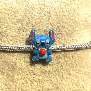 Peut inclure: Un charm en &eacute;mail bleu repr&eacute;sentant le personnage Stitch de Lilo & Stitch. Le charm a une langue rouge, des oreilles roses et est pos&eacute; sur une cha&icirc;ne argent&eacute;e. Un objet de collection.