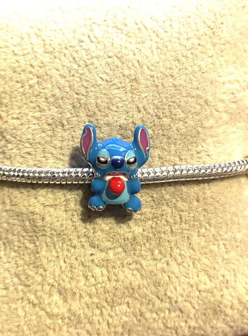 Peut inclure: Un charm en &eacute;mail bleu repr&eacute;sentant le personnage Stitch de Lilo & Stitch. Le charm a une langue rouge, des oreilles roses et est pos&eacute; sur une cha&icirc;ne argent&eacute;e. Un objet de collection.