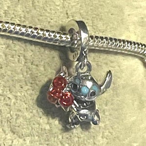 Pode incluir: Um berloque de prata com o personagem Stitch de Lilo & Stitch, segurando um cacho de bagas vermelhas. O berloque tem olhos azuis e est&aacute; preso a uma pulseira de prata.